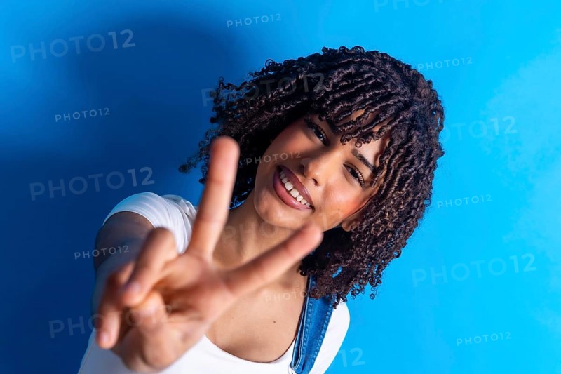 Cheerful young woman smiling and making a peace sign with both hands against a vibrant blue background, radiating joy and positivity