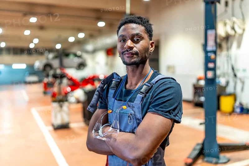 Young african american mechanic standing with crossed arms and protective eyewear in a car repair shop, exuding professionalism