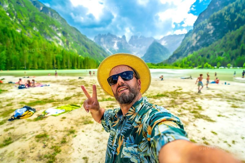 Tourist wearing sunglasses and straw hat gesturing peace sign while taking a selfie at the beach of lago di landro in the italian dolomites