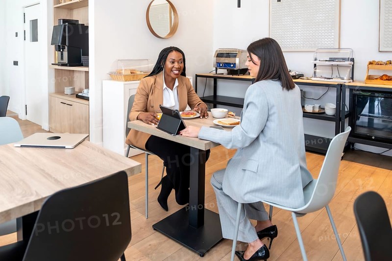 Two businesswomen are enjoying breakfast and discussing work using a digital tablet in a hotel restaurant