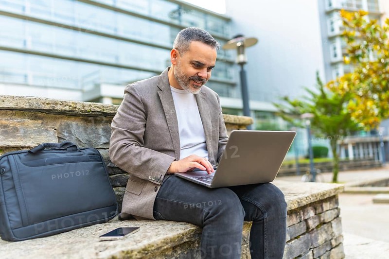 Happy mature caucasian businessman using laptop sitting on a stone bench outdoors in the financial district