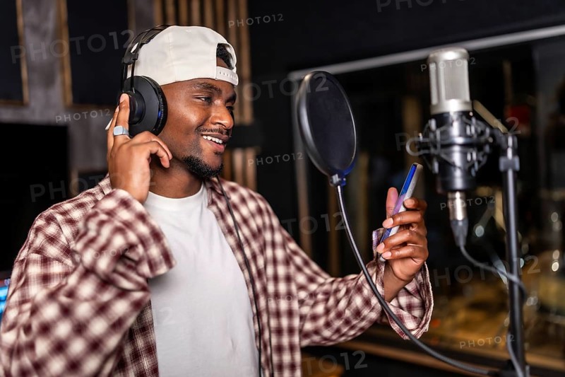 Young man wearing headphones and holding smartphone, recording a song or rap music in a professional studio