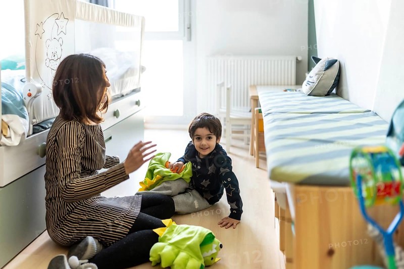 Mother and son are sitting on the floor, playing with a dinosaur costume in a brightly lit bedroom