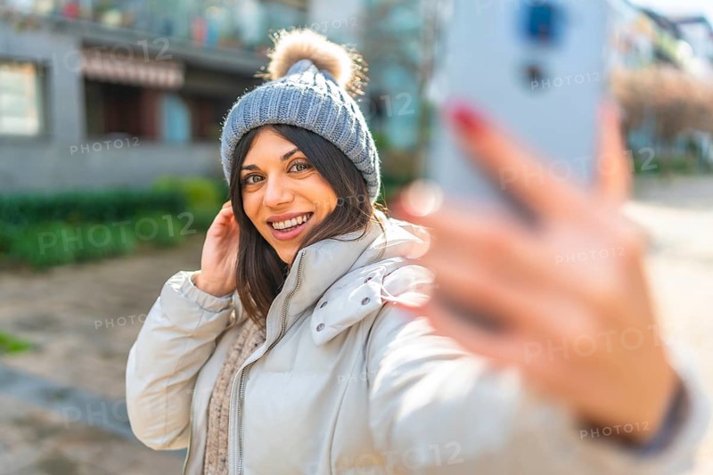 Beauty woman smiling while taking selfie with smartphone standing outdoors in a sun winter day