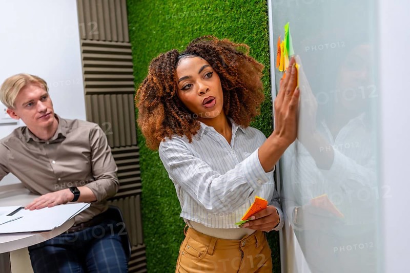 Businesswoman using colorful sticky notes while brainstorming new ideas on a whiteboard in a modern coworking space, with a male colleague sitting nearby