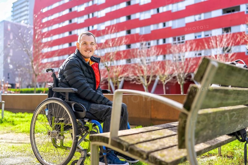 Portrait of a cheerful young disabled man sitting in a wheelchair enjoying a sunny day in a city park, promoting inclusivity and accessibility