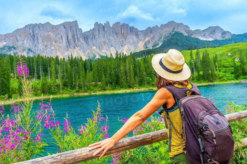 Female hiker with backpack enjoying the stunning turquoise waters of lake carezza with the latemar mountain range in the background, dolomites, italy