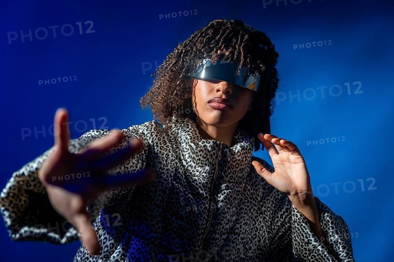 Stylish person with futuristic visor and patterned jacket extending hand on vibrant blue background