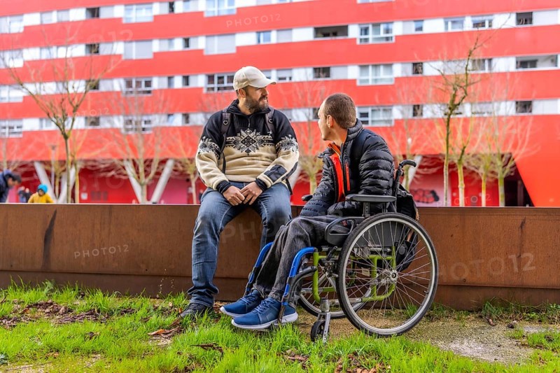 Two male friends are enjoying a conversation outside in the city, one of whom is using a wheelchair, demonstrating inclusion and friendship