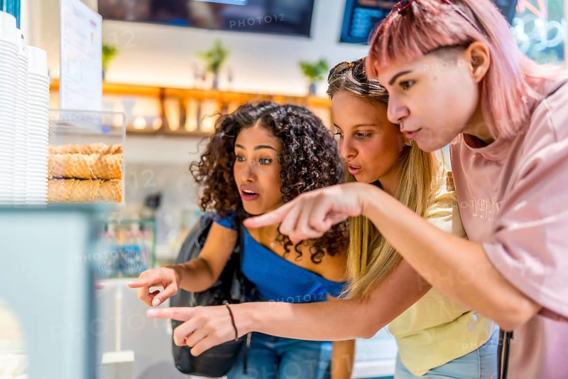 Side view of three diverse modern women pointing at ice cream glass counter choosing a flavor