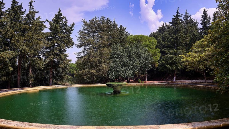A round lake with a central fountain surrounded by tall green trees under a blue sky with clouds, Italian Governor's Palace, Ruins, Italian colonial settlement, Eleousa, Rhodes, Dodecanese, Greek Islands, Greece, Europe