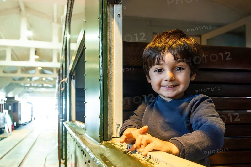 A young boy is smiling and looking out a window of a train. The train is in a station and the boy is sitting on a bench