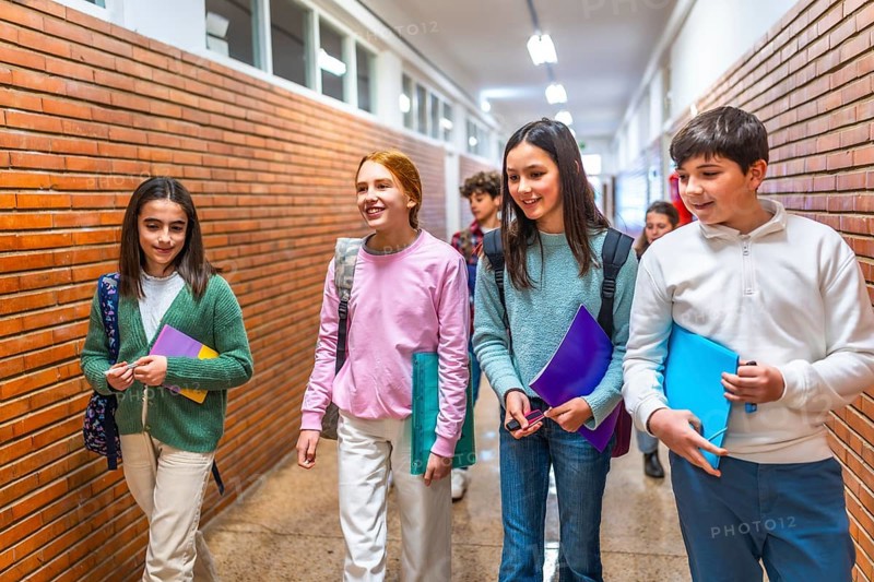 Cheerful students carrying books and folders are walking down school hallway between classes, chatting and smiling