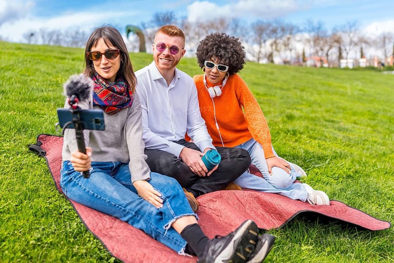 Three content creators sitting on a blanket in a park, recording a video blog using a smartphone with microphone