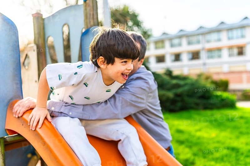 Grandfather holding grandson sliding down an orange slide at playground, having fun together on a sunny day