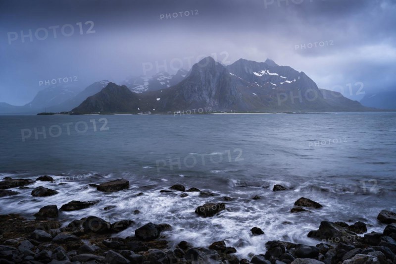Landscape with sea and mountains on the Lofoten Islands, view across the fjord to the small town of Flakstad and the mountain Flakstadtinden as well as other mountains. Rocks in the foreground. At night at the time of the midnight sun, clouds in the sky, bad weather. Long exposure, early summer. Flakstadoya, Lofoten, Norway, Europe