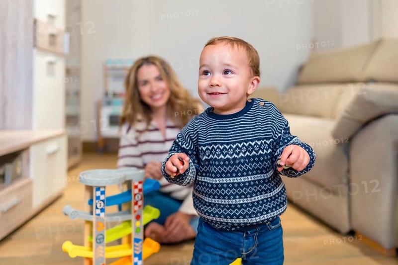 Happy baby boy learning to walk in living room with his mother watching and smiling, playing with a toy car ramp