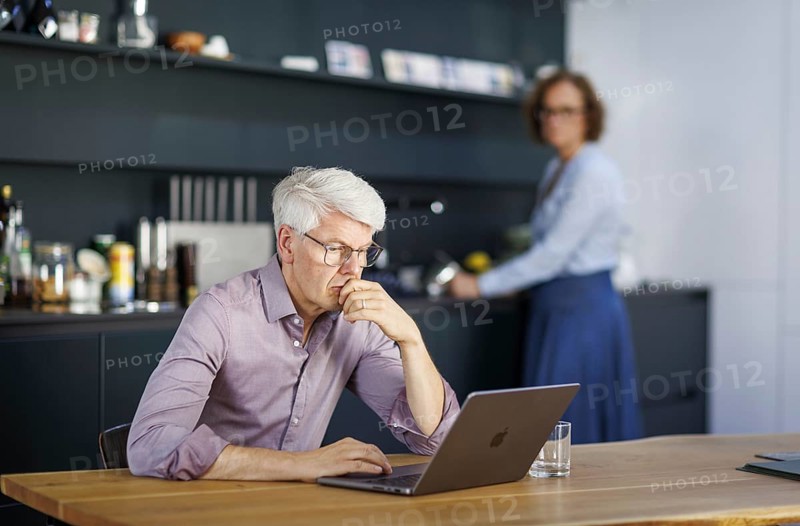 Symbolic photo on the subject of division of labour for couples in the household. A man sits in a kitchen at his laptop while a woman washes dishes in the background. Berlin, 13.08.2024