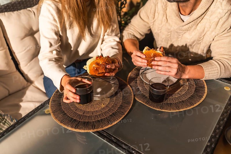 Couple sharing a relaxed moment, eating burgers and drinking beverages at a glass table, creating a cozy and intimate atmosphere