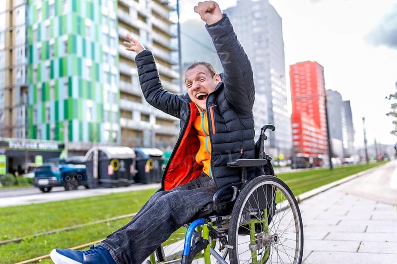 Young man in a wheelchair, raising his arms in celebration, embracing freedom and joy in a vibrant urban setting filled with modern architecture