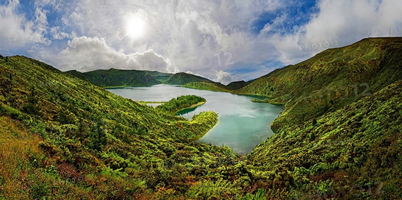 A clear lake surrounded by green hills and mountains under a blue sky with clouds on a sunny day, crater lake Lagoa do Fogo, Caldeiras, Sao Miguel Island, Azores, Portugal, Europe