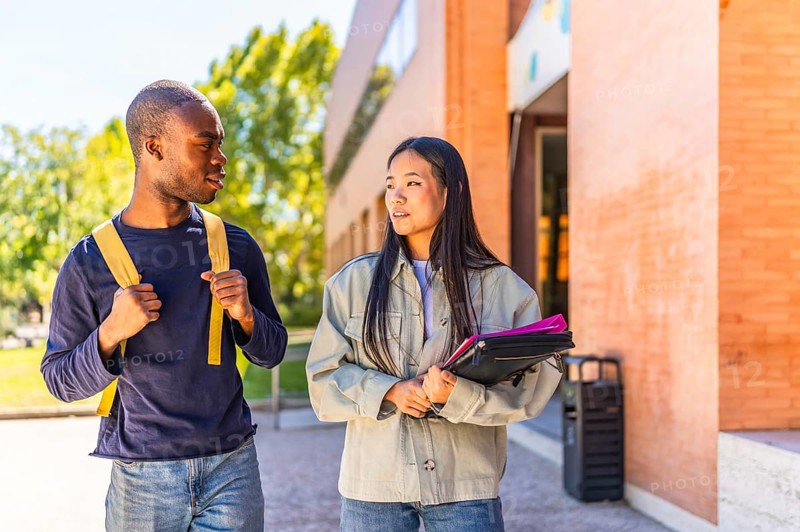 Two african and chinese multi-ethnic friends talking walking along university campus carrying folders and notebooks