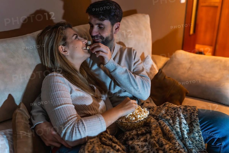 Couple enjoying a cozy night in, sharing popcorn and watching a movie together on the sofa