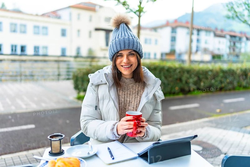 Beauty female caucasian entrepreneur working with digital tablet sitting in an outdoor cafeteria and warming hands with hot tea in a cold winter day
