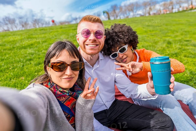 Three happy multi ethnic friends are taking a selfie in a park, gesturing victory signs and showing a reusable coffee cup