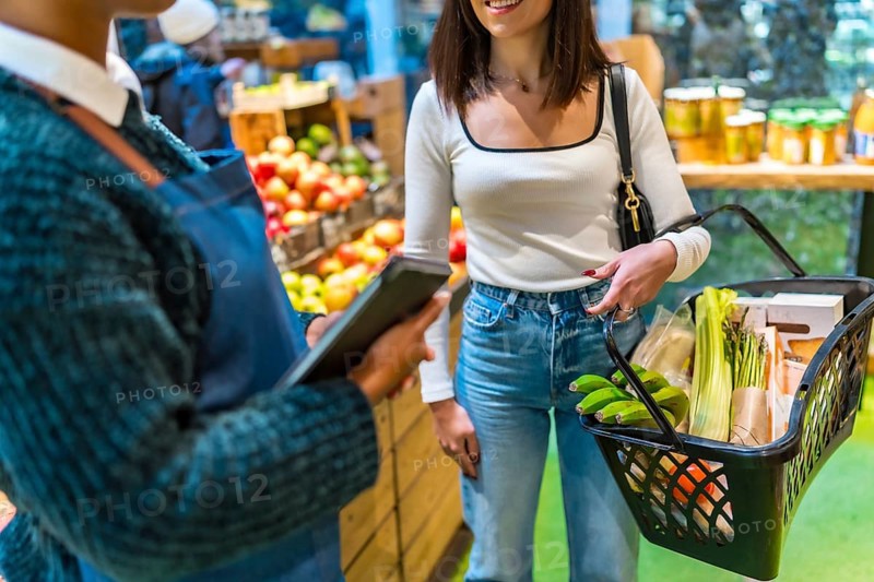 Saleswoman using digital tablet assisting customer carrying shopping basket with fresh produce in organic supermarket
