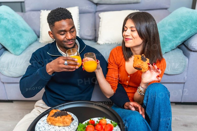 Multiethnic couple sits together, enjoying breakfast and wine, creating a cozy and intimate dining experience in their living room