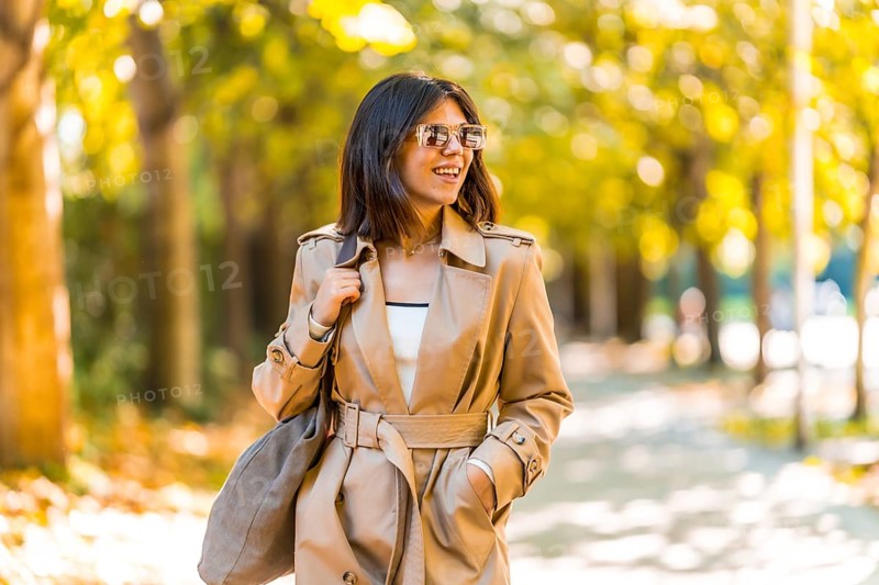 A woman wearing a tan trench coat and sunglasses walks down a sidewalk. She is smiling and she is enjoying her day. Autumn or winter concept in the city