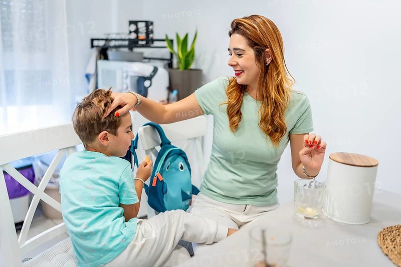 Tender caucasian mother sitting on the kitchen stroking her son's head before he goes to school