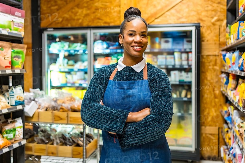 Smiling young african american saleswoman wearing an apron, standing confidently with arms crossed in a grocery store aisle filled with organic food