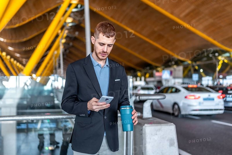 Young businessman using smartphone and holding reusable coffee cup while waiting for taxi or rideshare at airport