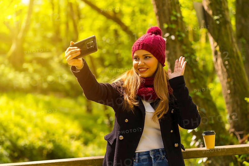 Young man in a black jacket, scarf and red woolen hat enjoying in an autumn park, waving on a video call with the phone
