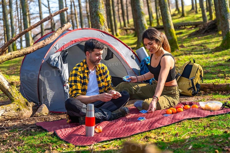 Hikers playing cards near their tent in the forest, enjoying a camping trip with fruits and snacks