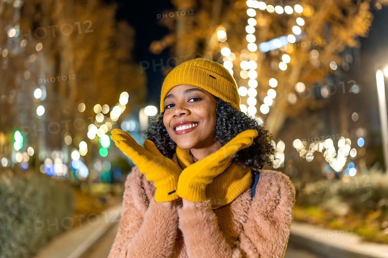 Portrait of a cheerful young woman wearing yellow woolen gloves and hat enjoying christmas lights in the city at night