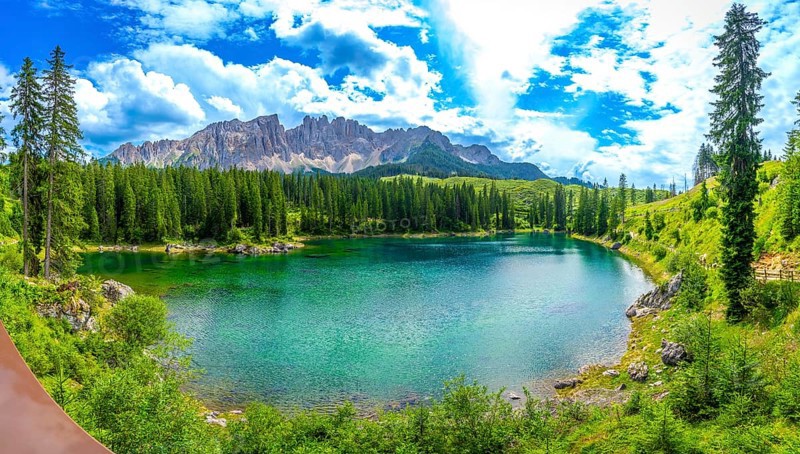 Emerald green water of lake carezza reflecting latemar mountain range in italian dolomites, surrounded by coniferous forest on a sunny summer day