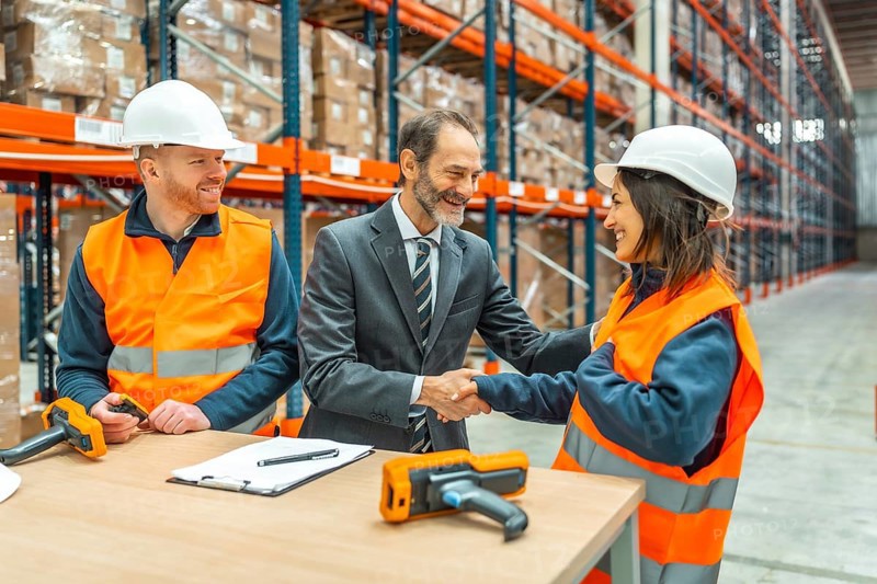 Manager shaking hands with a smiling female worker in a logistics warehouse, a male worker is looking at them, barcode readers and contract on the table