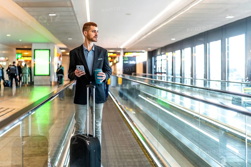Businessman is walking on moving walkway at airport while using smartphone and holding coffee