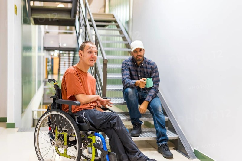 Man with cerebral palsy talking with a work colleague indoors in a modern coworking with staircase