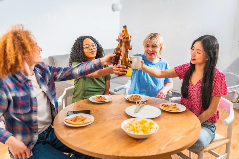 Four diverse friends are enjoying a meal together, toasting drinks and eating pizza at a round wooden table