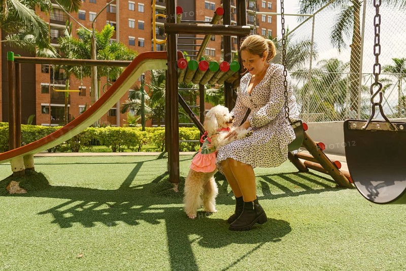 Happy woman in a stylish dress swinging on a playground swing, joyfully playing with her small white dog dressed in pink, creating moments of fun and affection in the sunny park