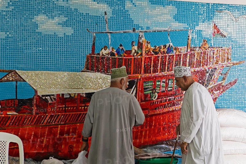 Omani men in dishdashas in front of the wall mosaic of a ship in Muttrah Fish Market, Muscat, Oman, Asia