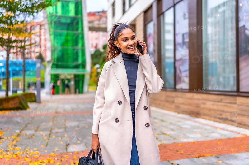 African businesswoman walking while talking to the mobile phone along a city street with fallen leaves in autumn