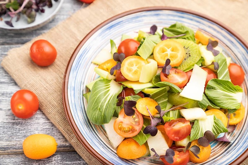 Vegetarian salad of pac choi cabbage, kiwi, tomatoes, kumquat, microgreen sprouts on gray wooden background and linen textile. Top view, close up, selective focus