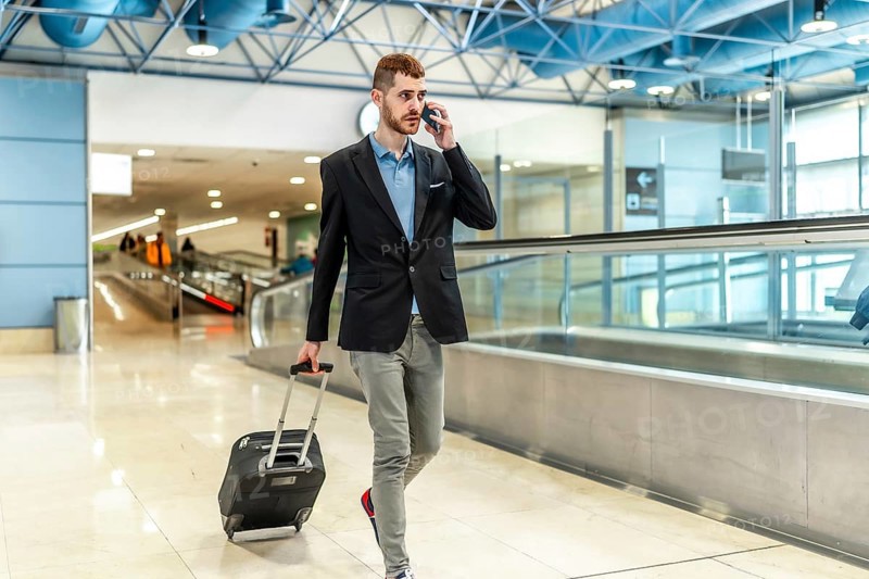 Businessman pulling a suitcase while talking on the phone, confidently walking through a bustling airport terminal