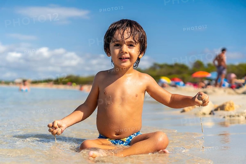 Happy child playing with sand sitting on the shore of a paradisiacal beach with crystalline waters and other tourists in the background