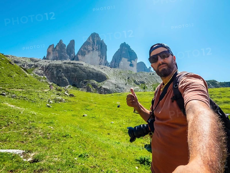 Photographer taking a selfie while hiking showing thumbs up sign with tre cime di lavaredo mountains in the background